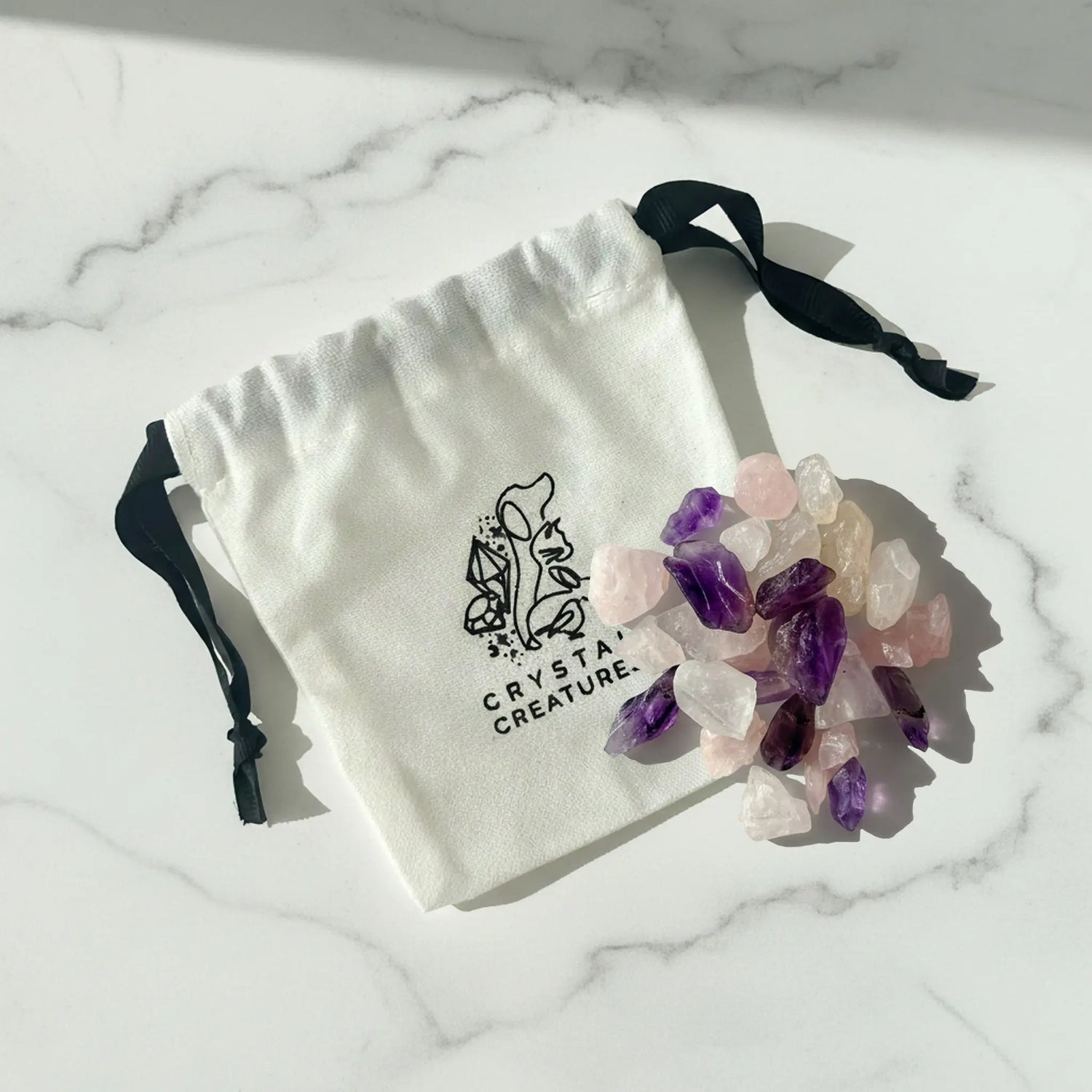 White drawstring bag with 'Crystal Creature' logo containing crystals on a marble surface.