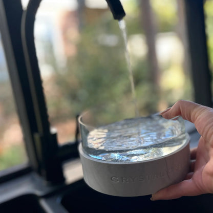 Hand holding a Crystal Elixir Pet Water Bowl  with water being poured into it, blurred background of trees.